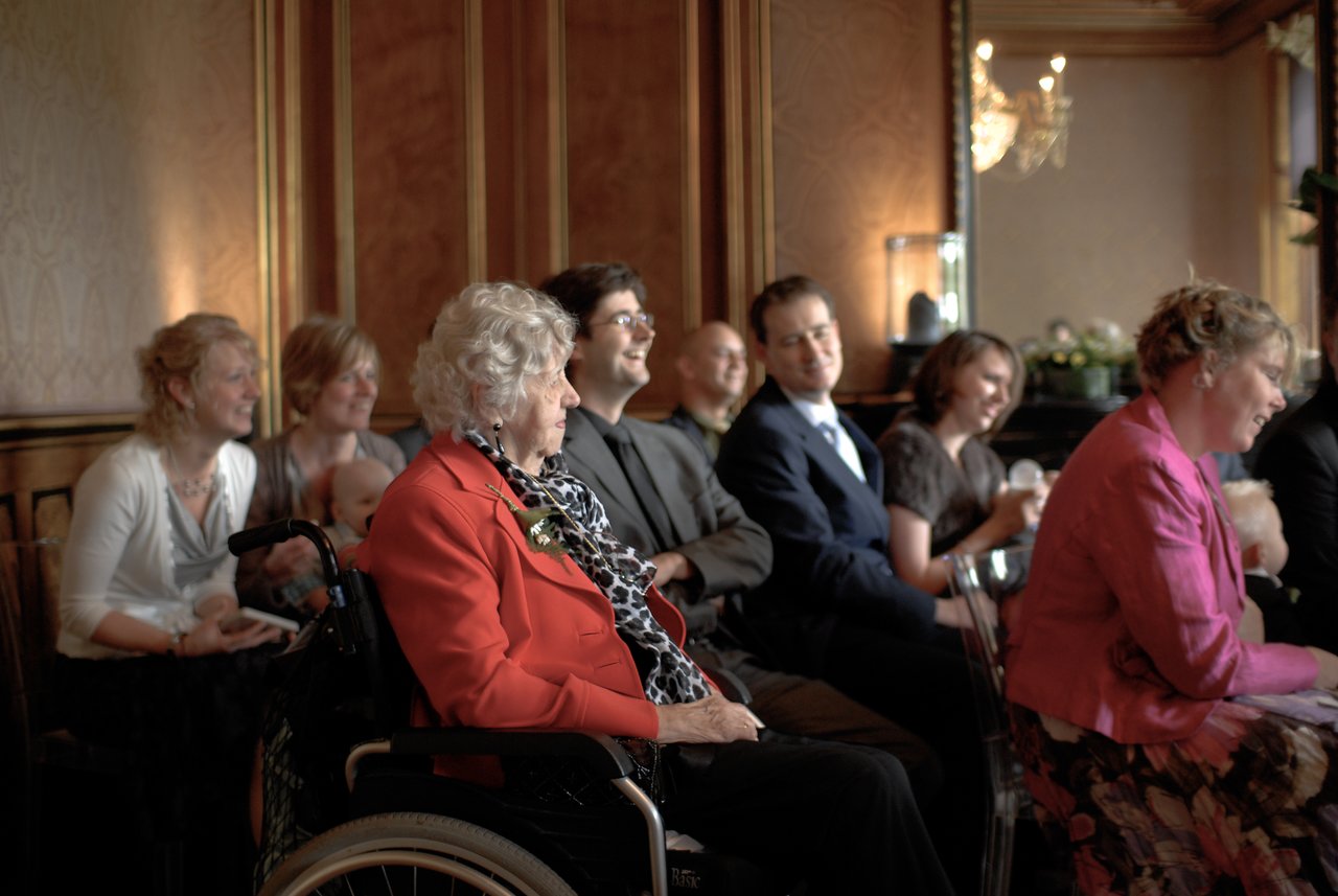 Guests seated at a wedding, including an elderly woman in a wheelchair, smiling and enjoying the ceremony.