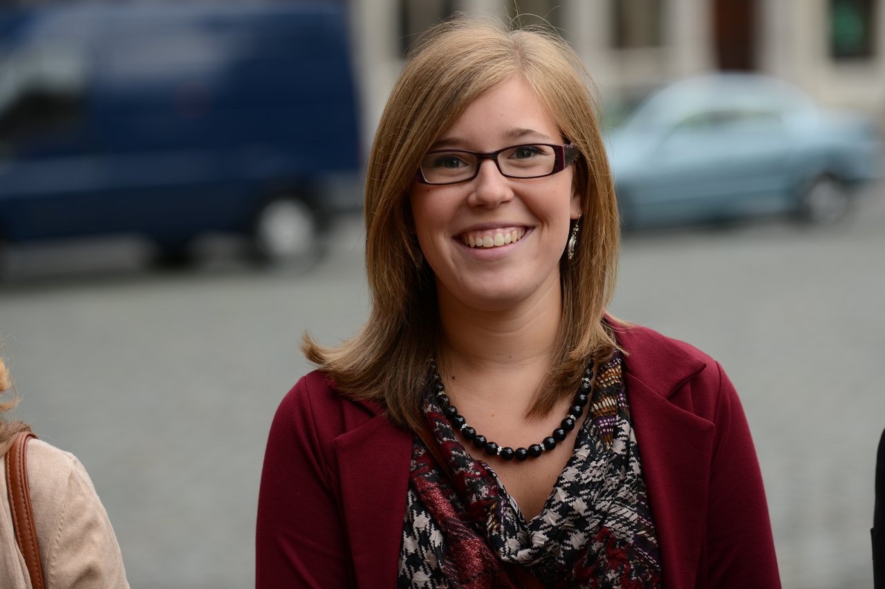 A smiling woman wearing glasses and a burgundy jacket stands outdoors, looking at the camera.