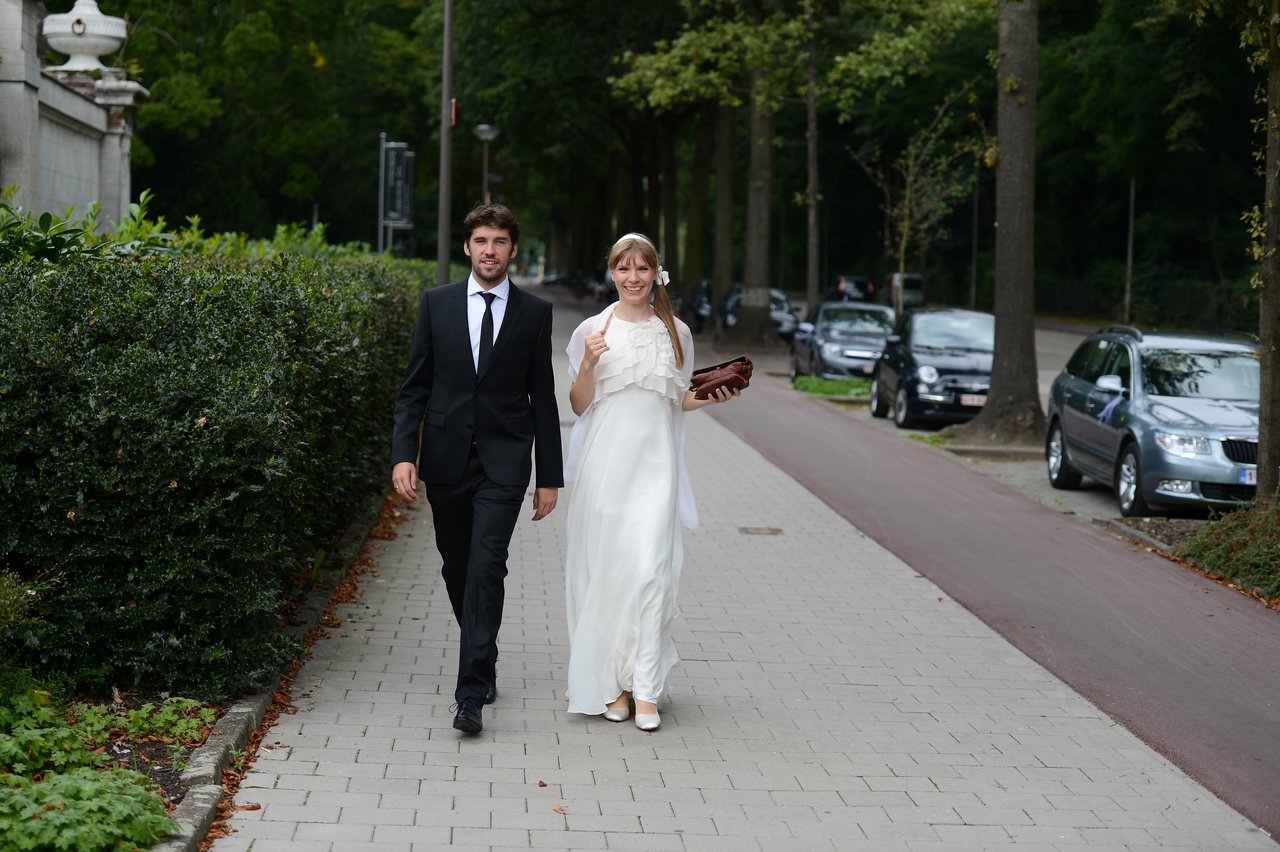 A bride and groom walk together on a paved path, dressed in wedding attire, smiling.