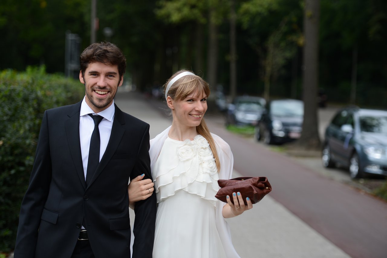 A smiling bride and groom walk arm in arm outdoors, dressed in wedding attire.