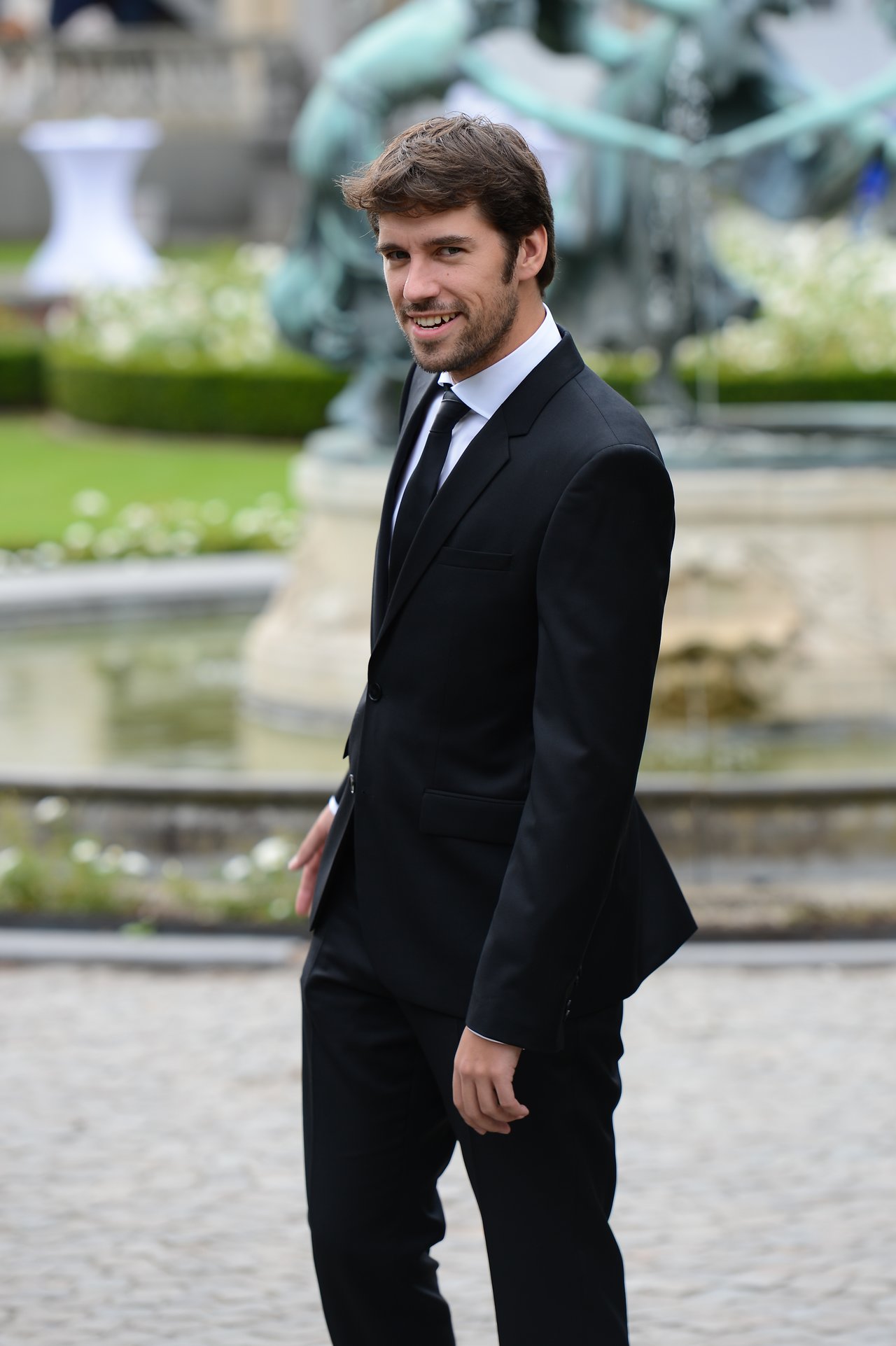 A man in a black suit smiles while walking outdoors at a wedding venue with a fountain in the background.