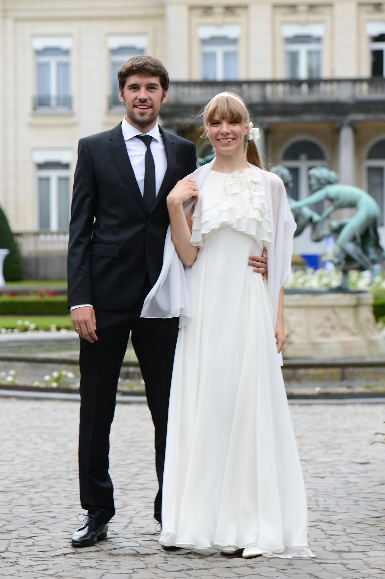 A bride and groom stand together, smiling at the camera in formal wedding attire outside a grand building.