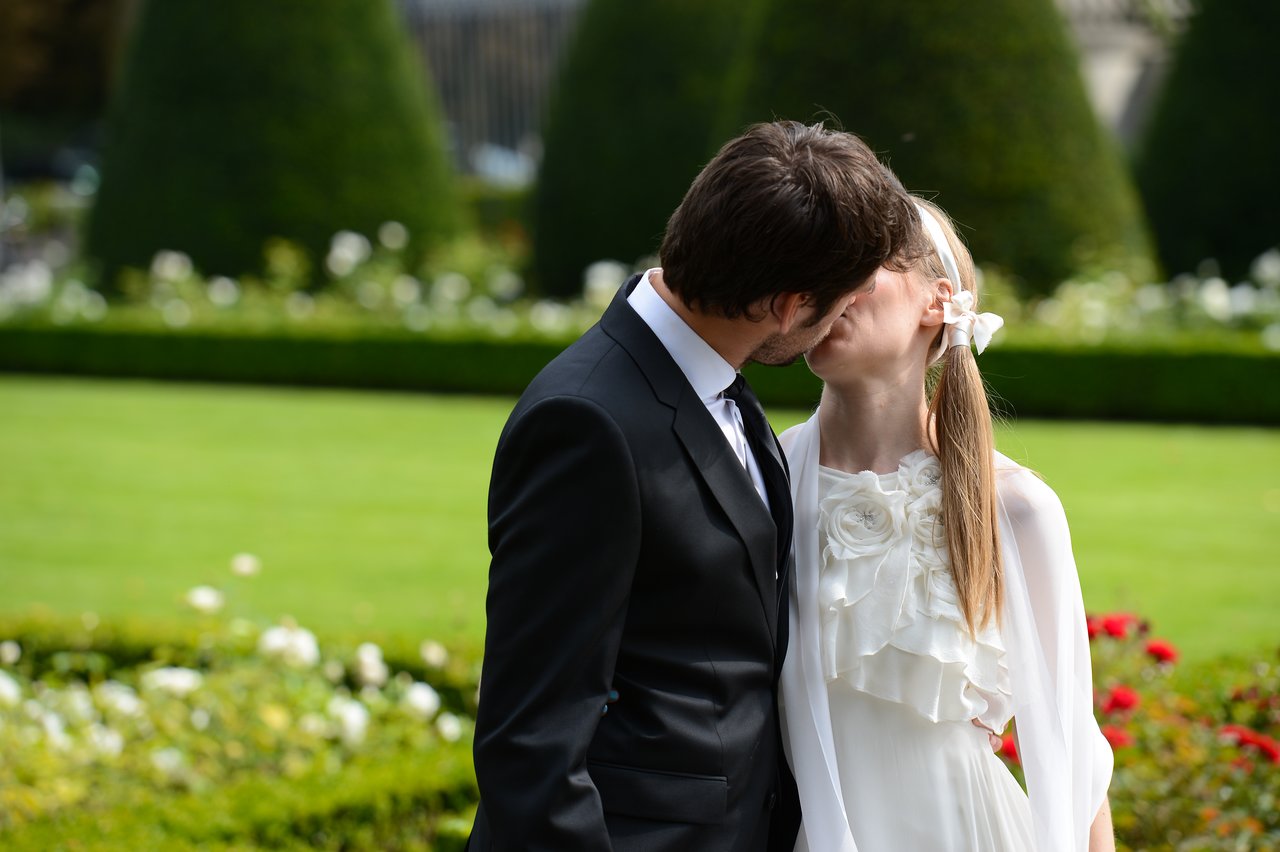 A newlywed couple shares a kiss in a garden, dressed in wedding attire.