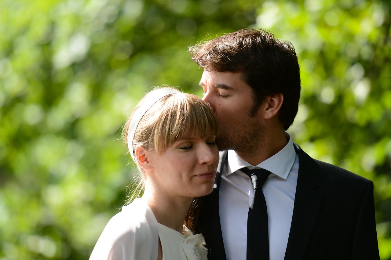 A groom in a suit kisses his bride on the forehead as she closes her eyes and smiles softly.