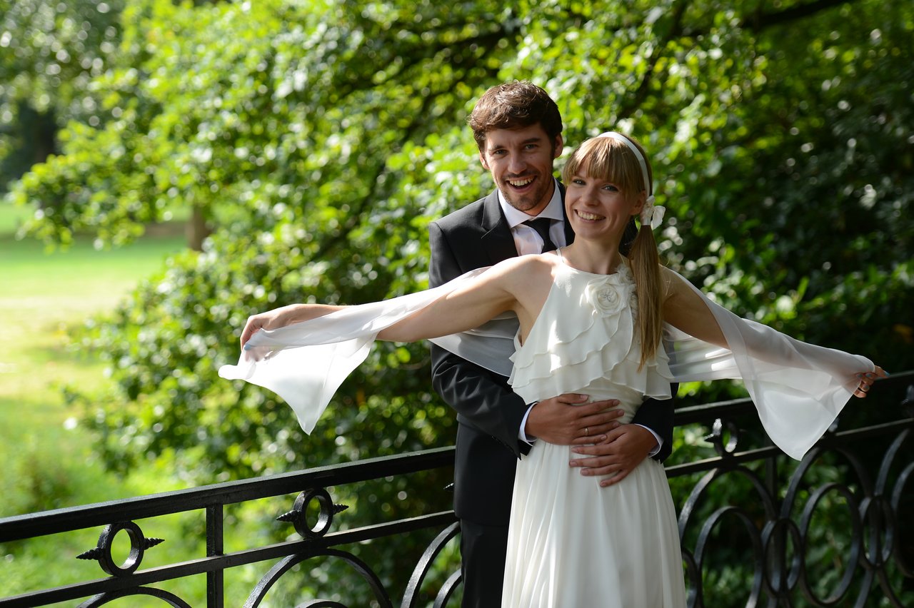 A bride and groom stand on a bridge, smiling as he embraces her from behind.