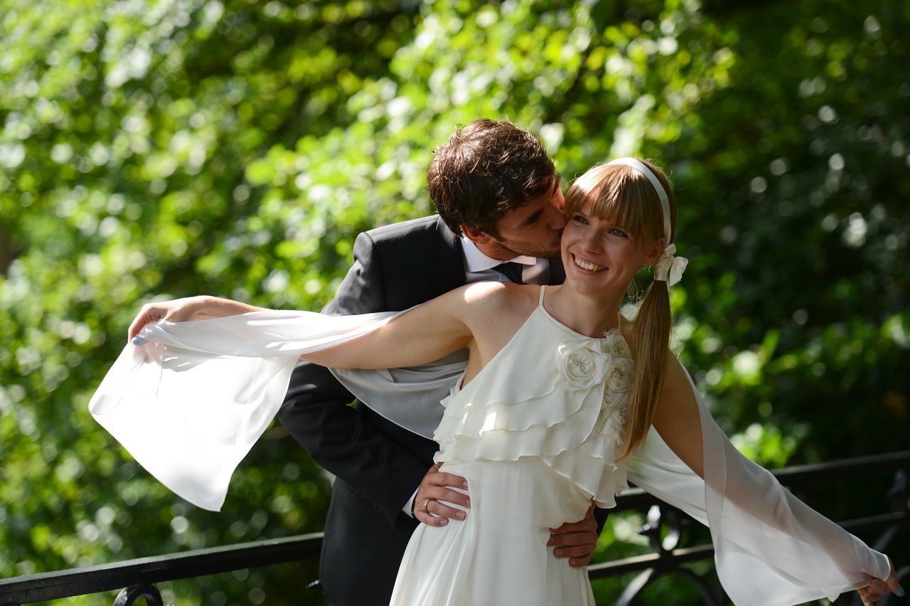 A groom in a suit kisses his smiling bride on the cheek as she spreads her arms in joy.