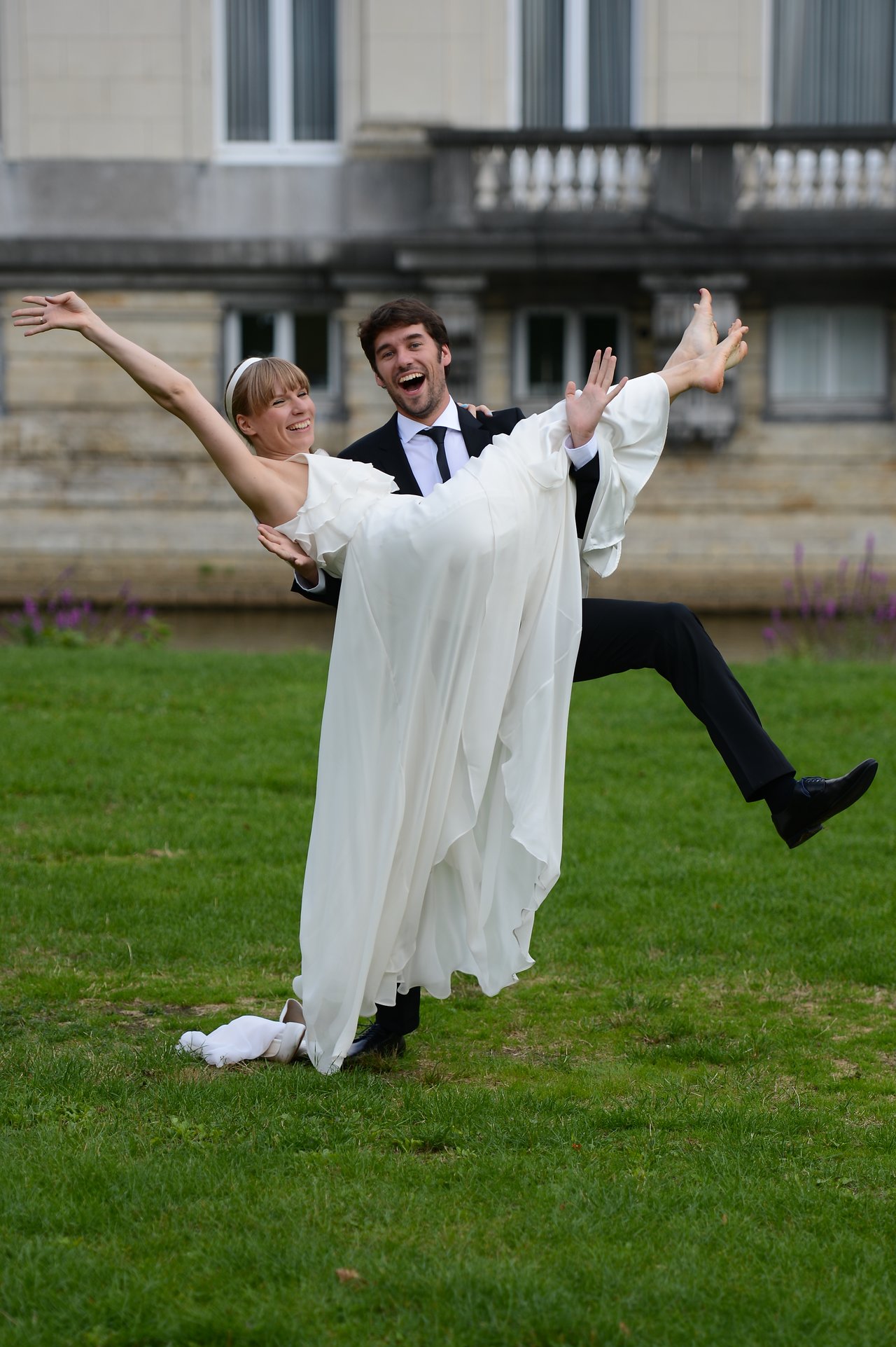 A groom in a suit playfully lifts the bride in a white dress as they smile outdoors.