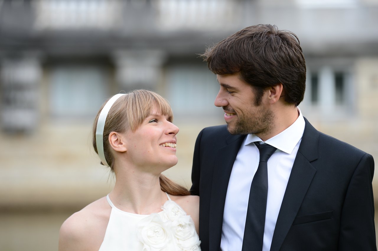 A bride and groom smile at each other, dressed in wedding attire, standing outdoors.