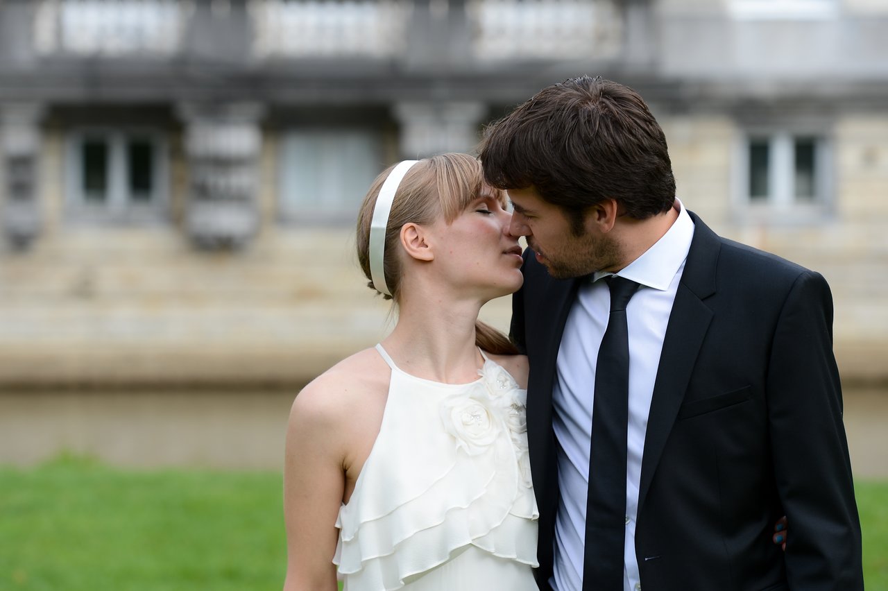 A bride and groom stand close together outdoors, leaning in for a kiss.