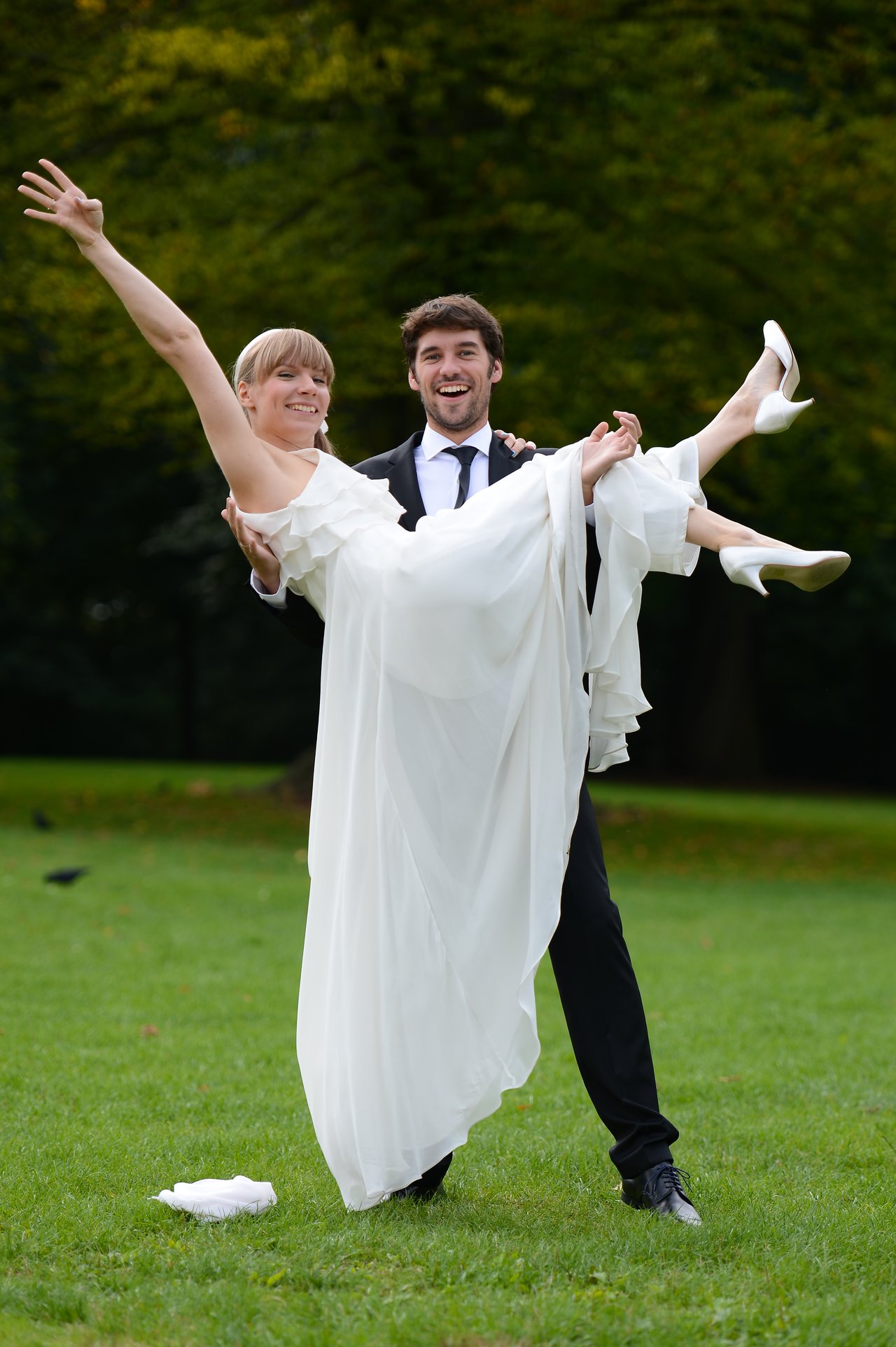 A groom in a suit joyfully lifts his bride in a white wedding dress as they smile outdoors.