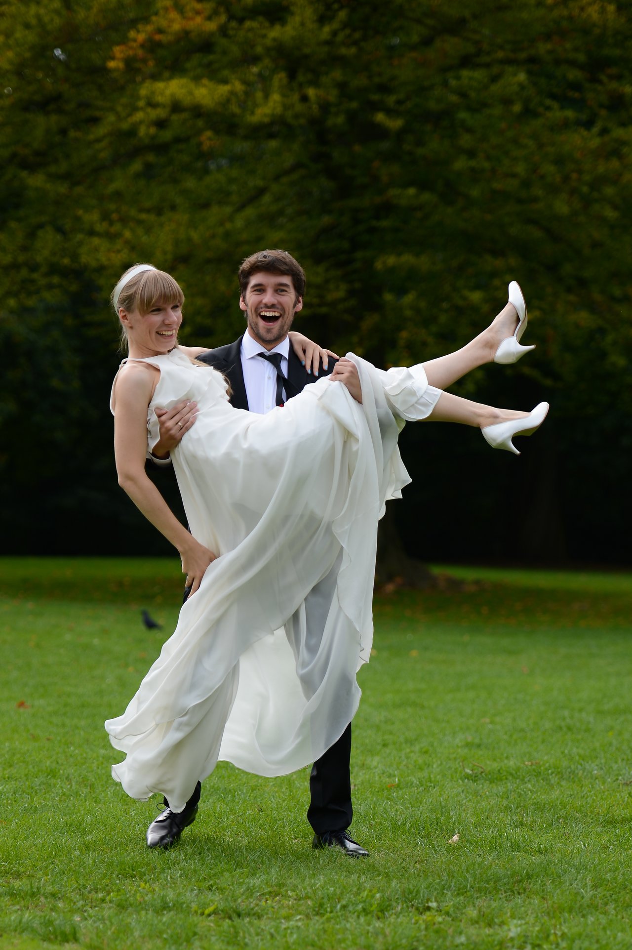 A groom in a suit joyfully lifts his bride in a white dress while smiling in a grassy park.