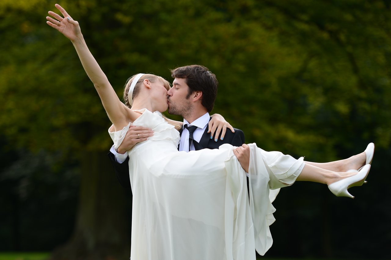 A groom in a suit carries his bride in a white dress as they kiss, celebrating their wedding day.