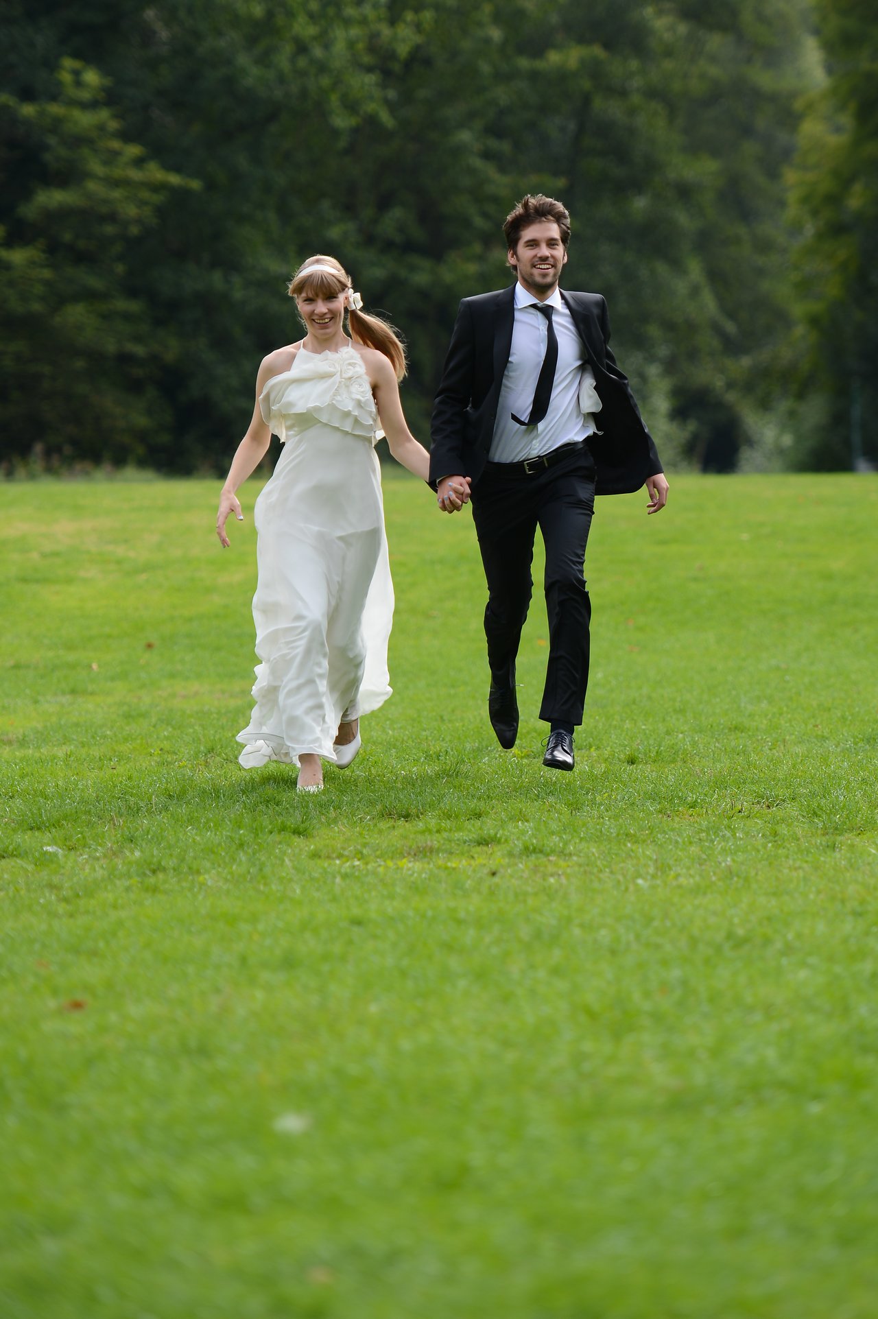 A bride and groom hold hands and run across a grassy field, smiling.