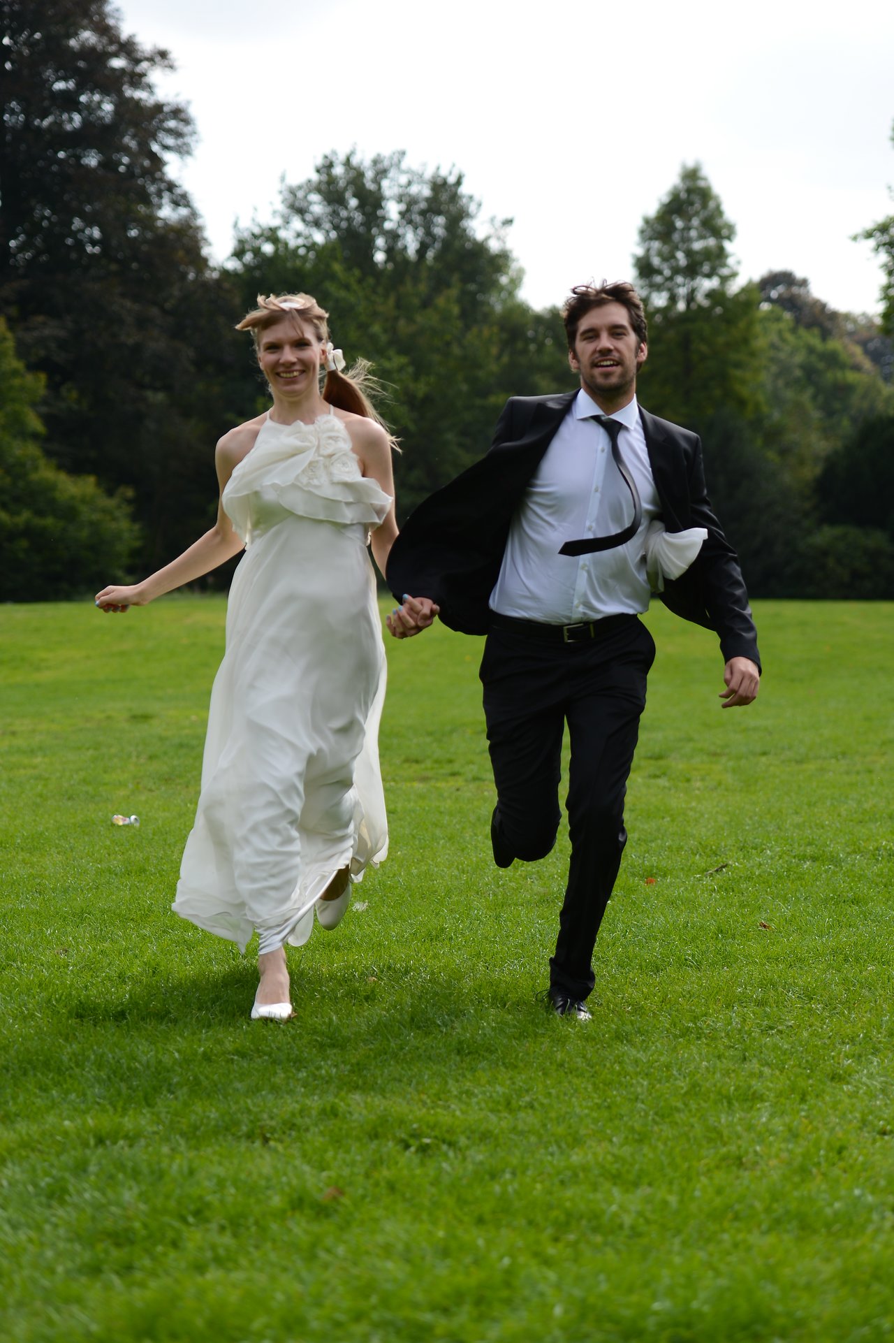 A bride and groom run hand in hand across a grassy field, smiling.