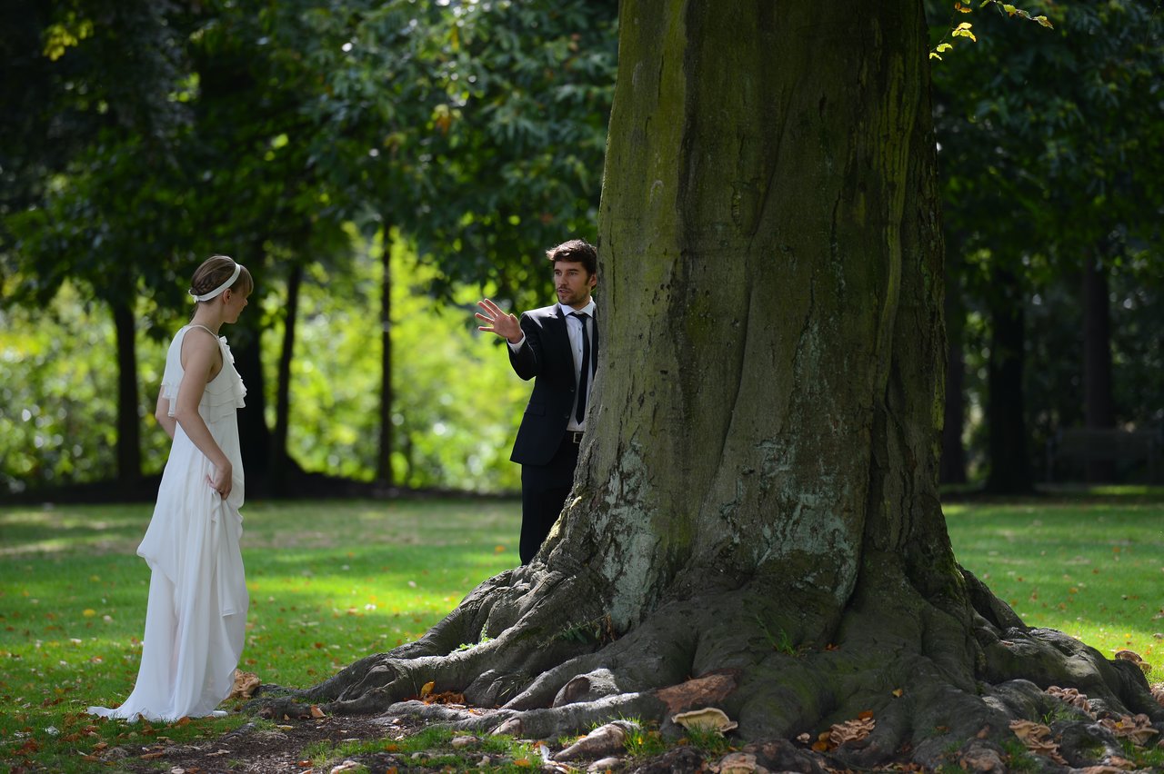 A bride and groom stand near a large tree in a park, looking at each other.