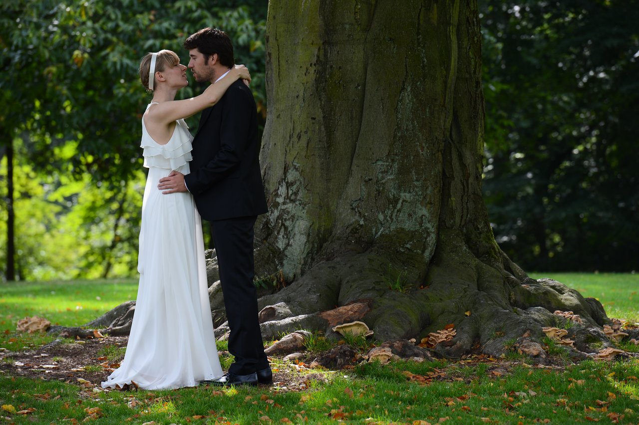 A bride and groom stand close together under a large tree, embracing and looking into each other's eyes.