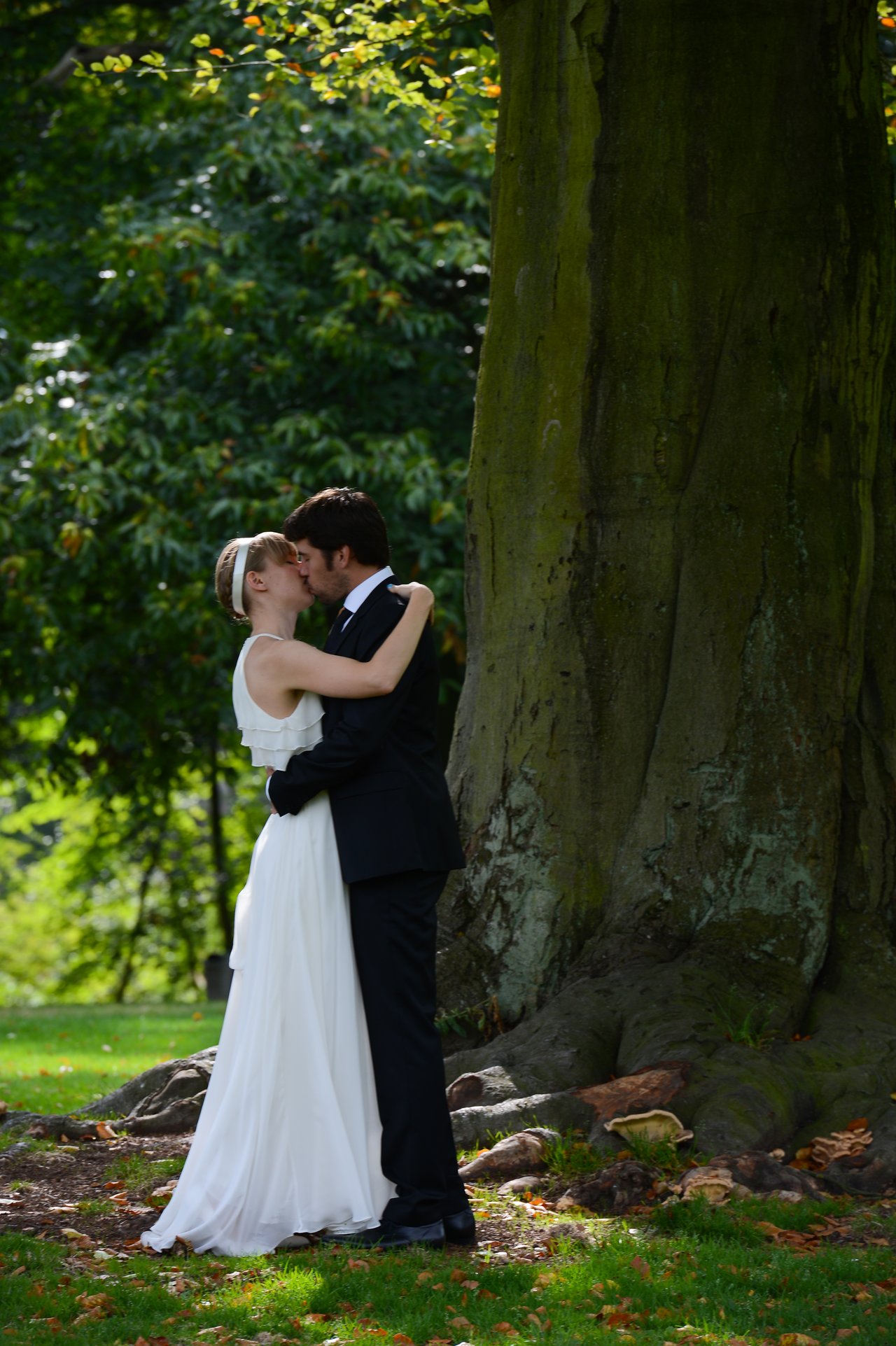 A bride and groom embrace and kiss outdoors near a large tree on their wedding day.