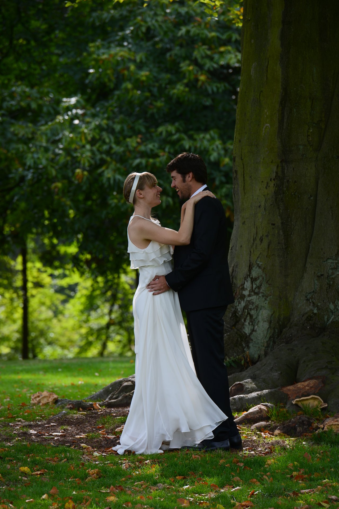 A bride and groom stand close together outdoors, smiling and embracing near a large tree.