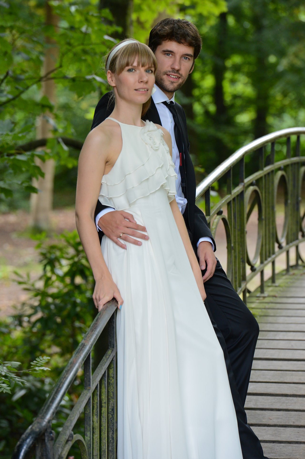 A bride and groom stand together on a bridge, with the groom's arm around the bride's waist.