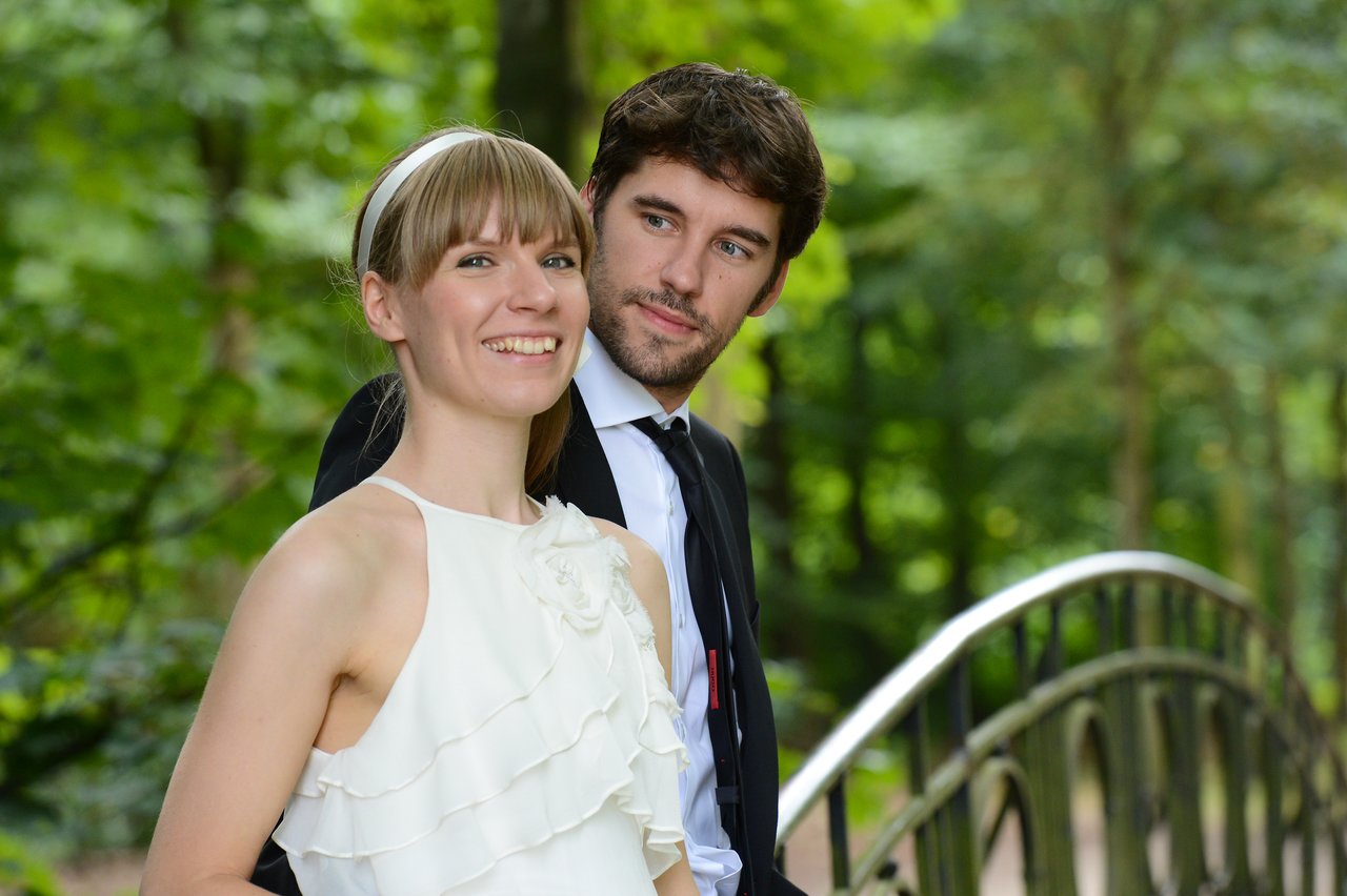A bride and groom stand together on a bridge, smiling in an outdoor setting.