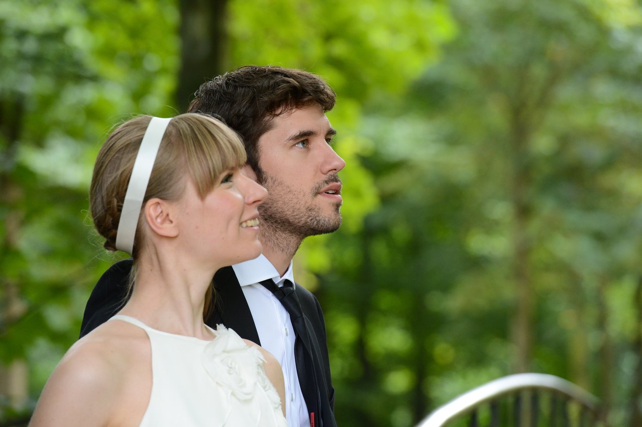 A bride and groom stand together outdoors, looking ahead with happy expressions.