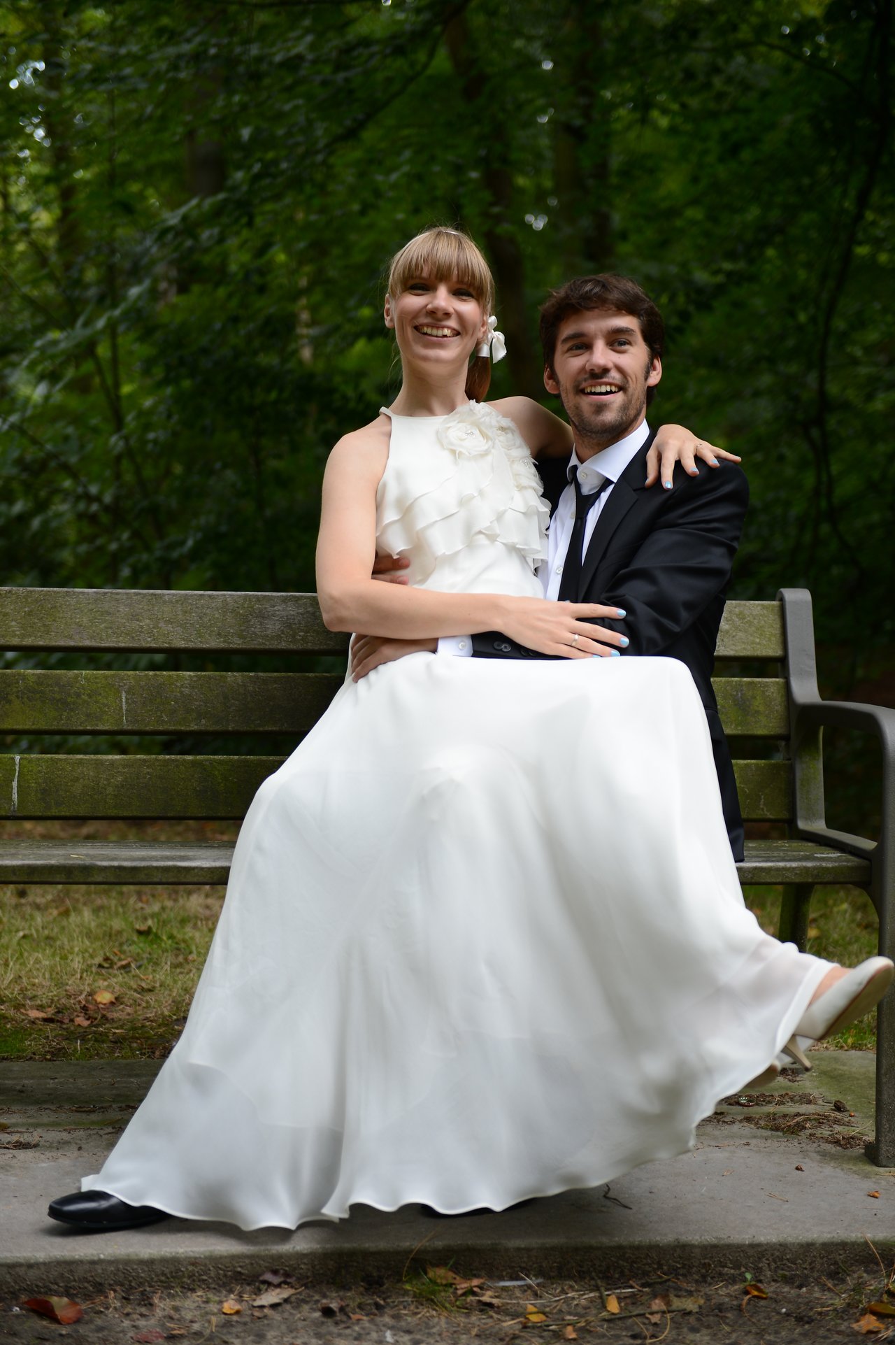A bride in a white dress sits on a groom's lap on a bench, both smiling.