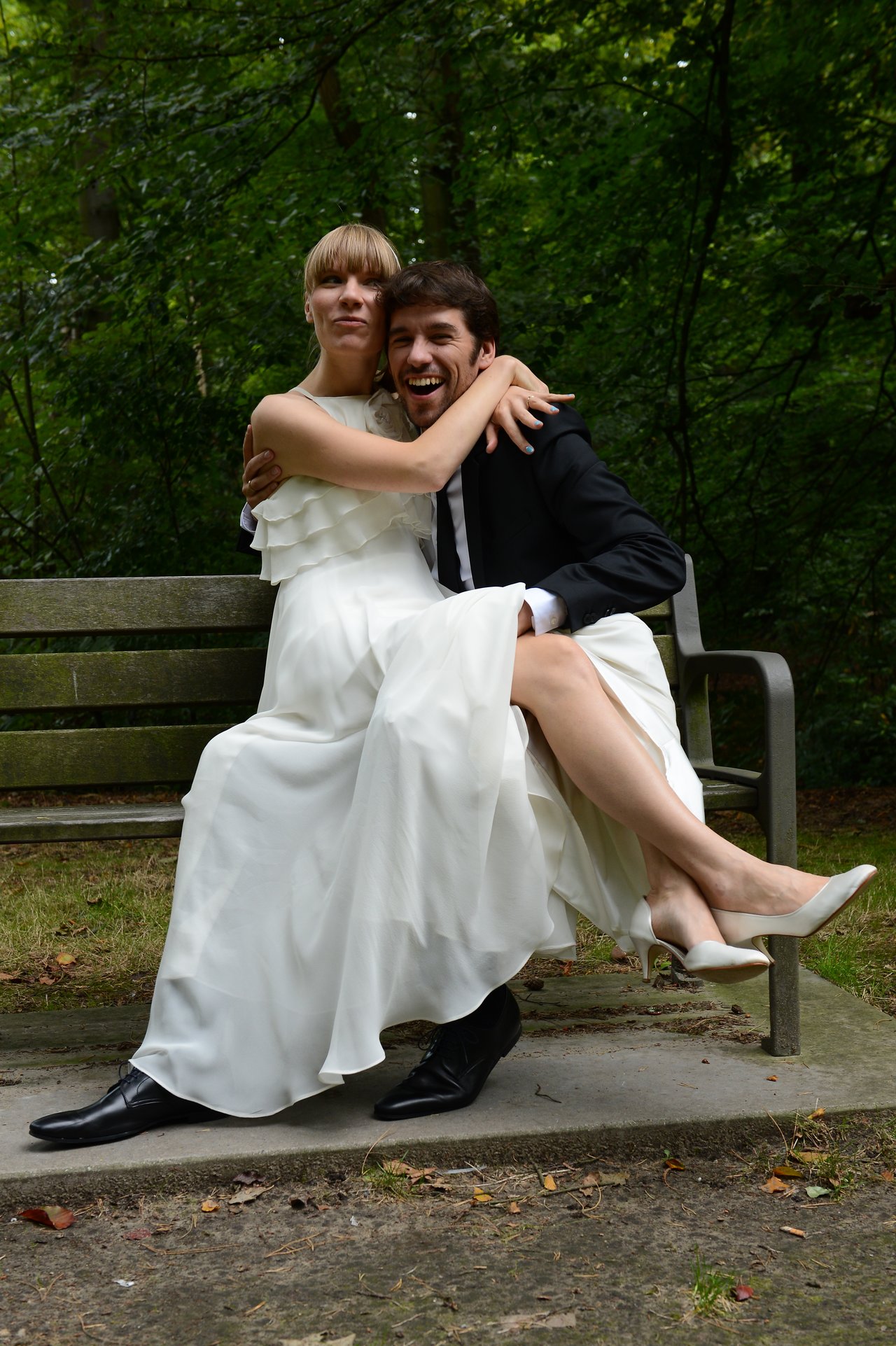 A newlywed couple in wedding attire sits on a bench, smiling and embracing each other.