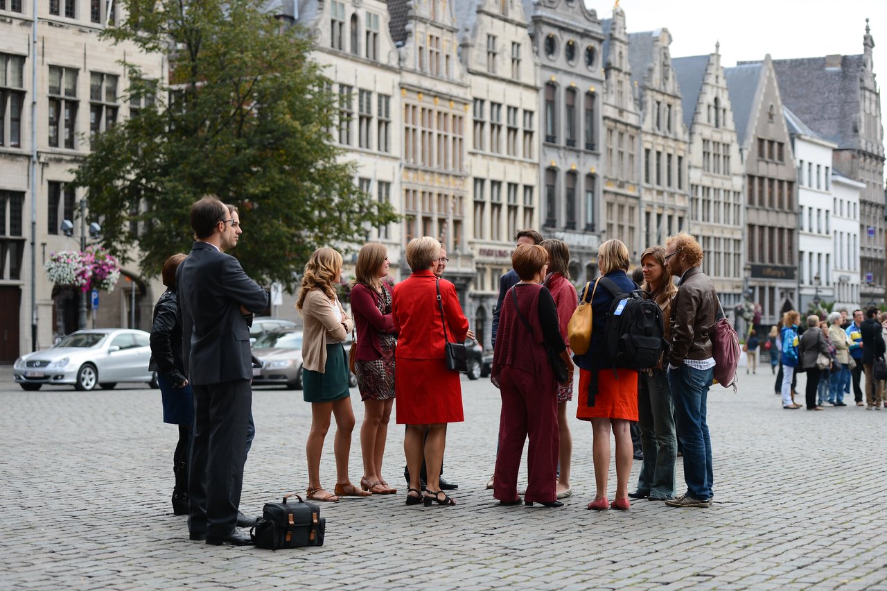 A group of people in formal and casual attire stand in a public square, engaged in conversation.