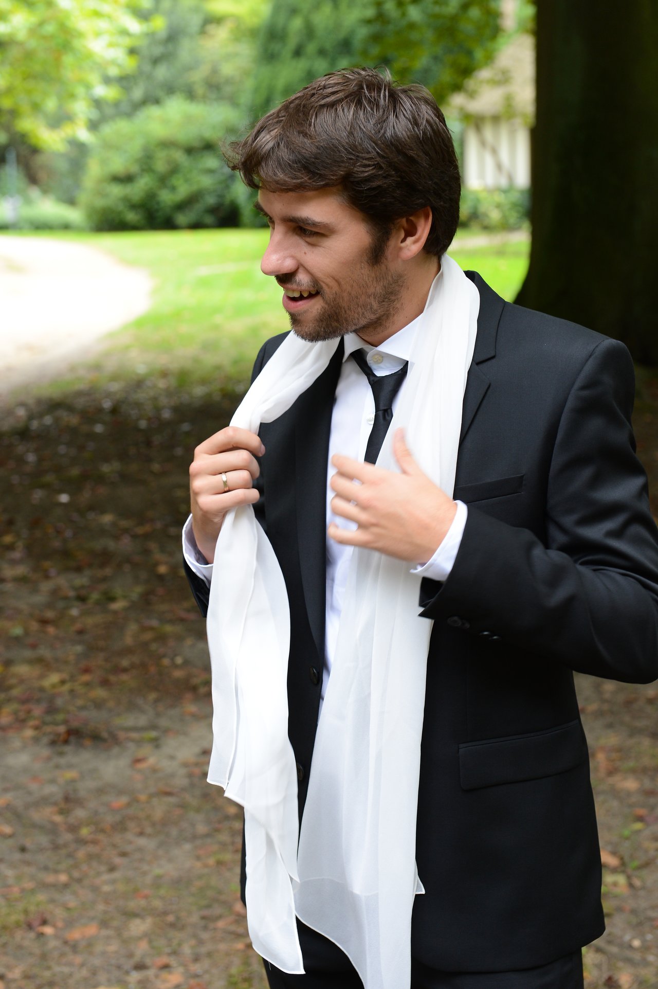 A man in a suit adjusts a white scarf while smiling outdoors during a wedding.