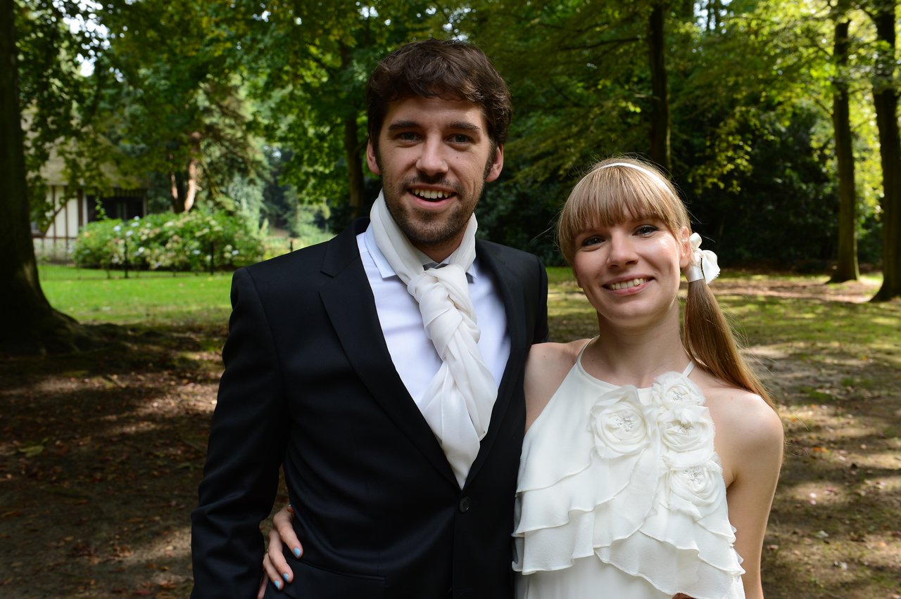 A smiling couple in wedding attire poses together outdoors, looking at the camera.