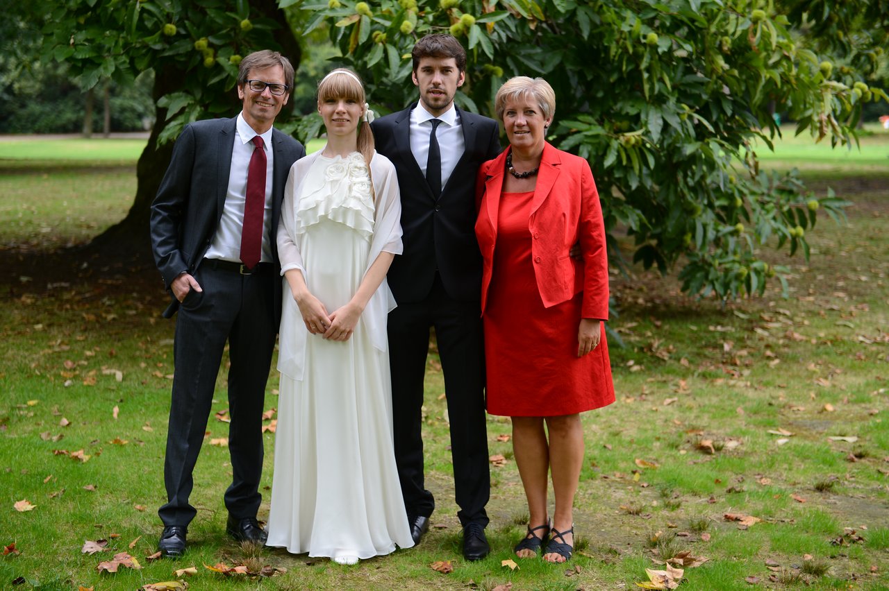 A bride and groom stand with two family members outdoors, dressed in formal attire for a wedding.
