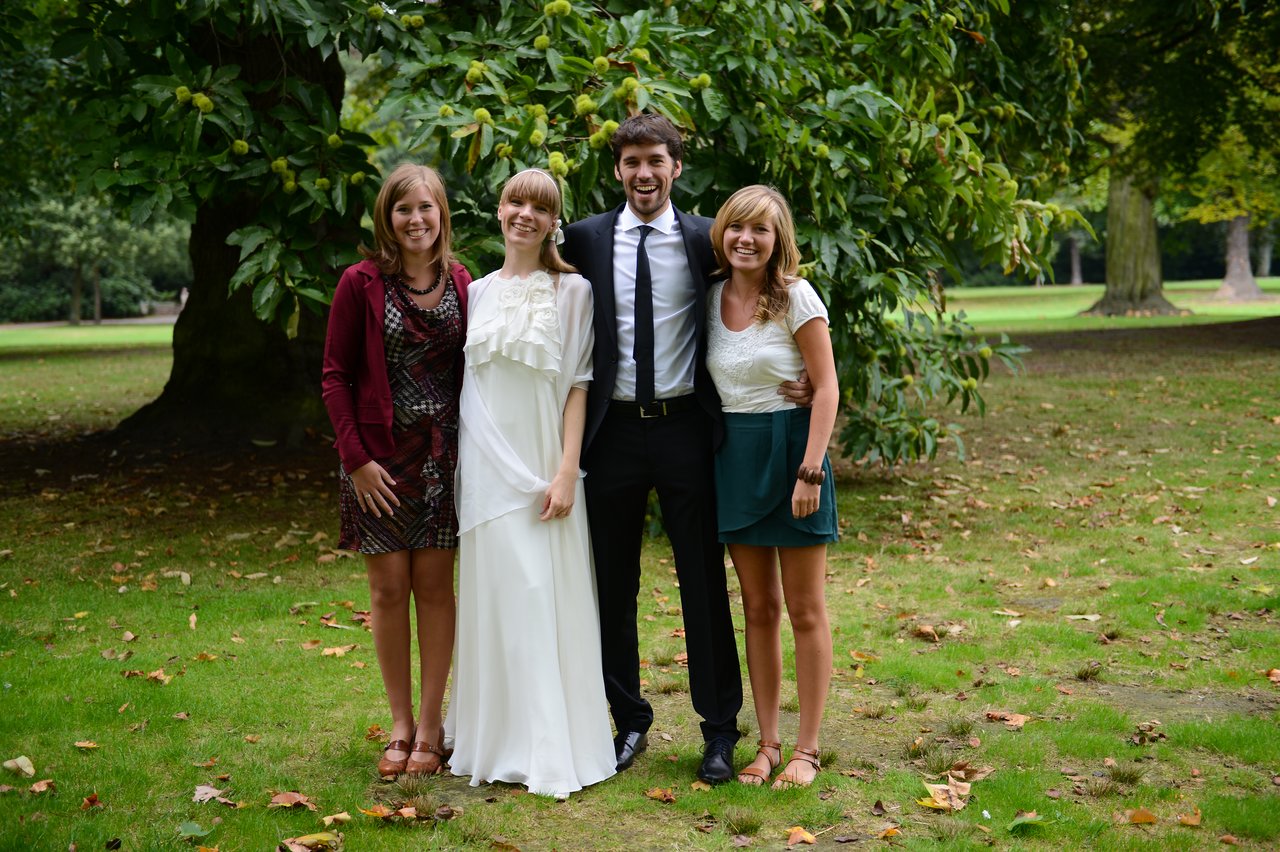 Bride and groom stand smiling with two friends in a park, dressed in formal attire for their wedding.
