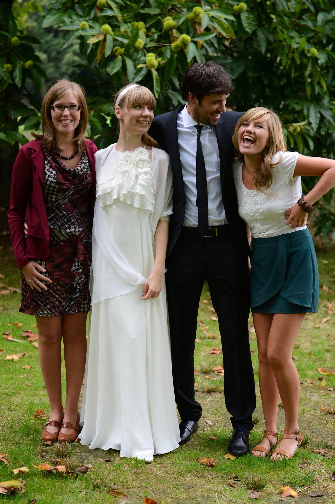 A bride and groom stand outdoors with two friends, all smiling and laughing together.