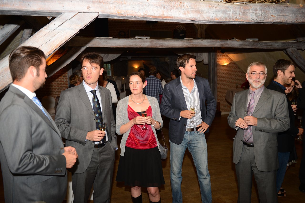 A group of wedding guests in formal attire stand together, holding drinks and talking in a warmly lit venue.