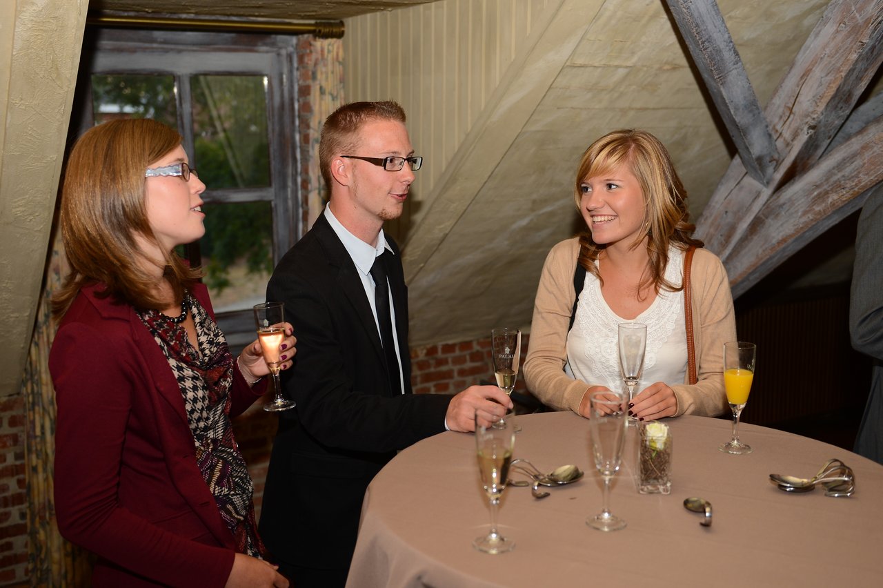 Three guests stand around a table, holding drinks and talking at a wedding reception.