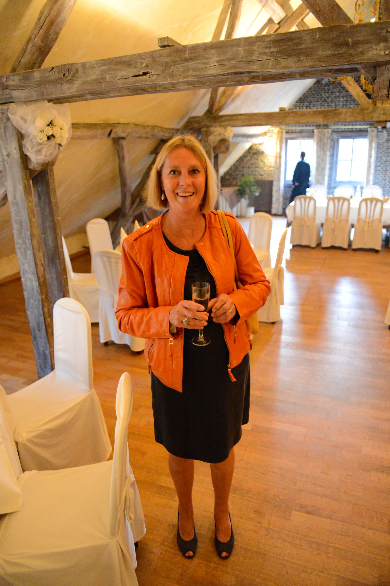 A woman in a black dress and orange jacket smiles while holding a glass of champagne at a wedding venue.
