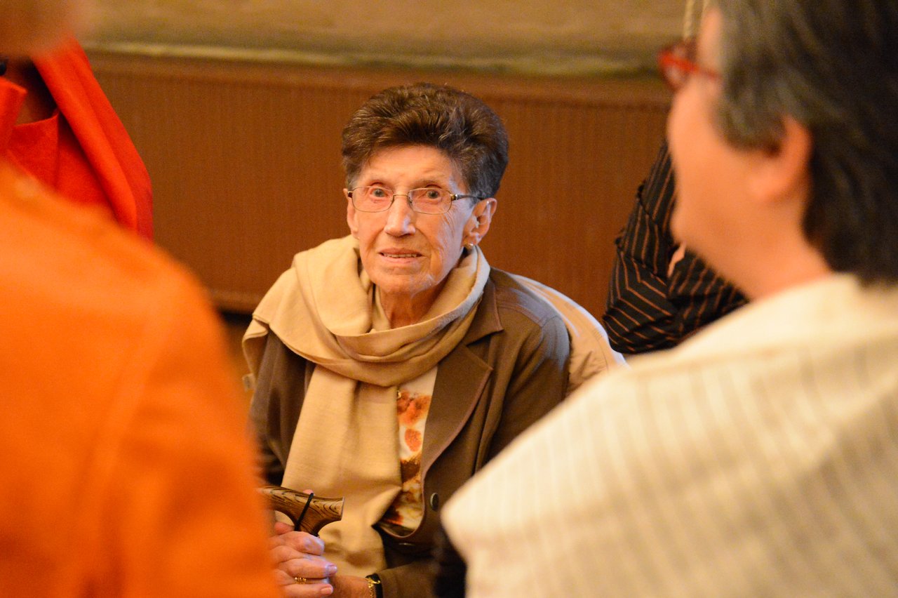 An elderly woman with glasses sits and listens while surrounded by people at a wedding gathering.