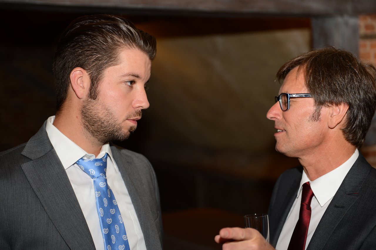 Two men in suits engaged in conversation at a wedding reception, one holding a glass.