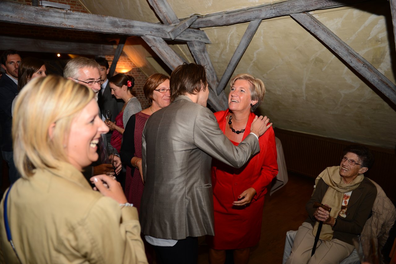 A man in a suit greets a smiling woman in red at a wedding reception with guests around them.