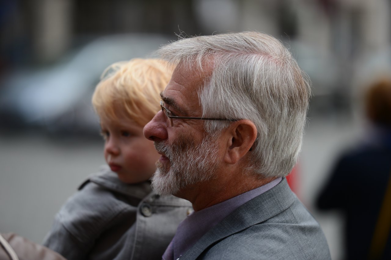 An older man in a suit holds a young child in a coat, both looking in the same direction.