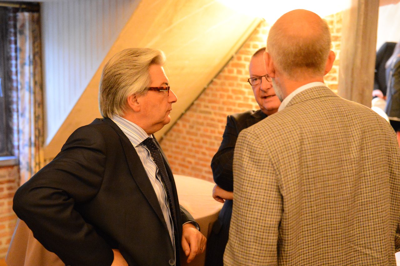 Three men in suits stand close together, engaged in conversation at an indoor wedding reception.