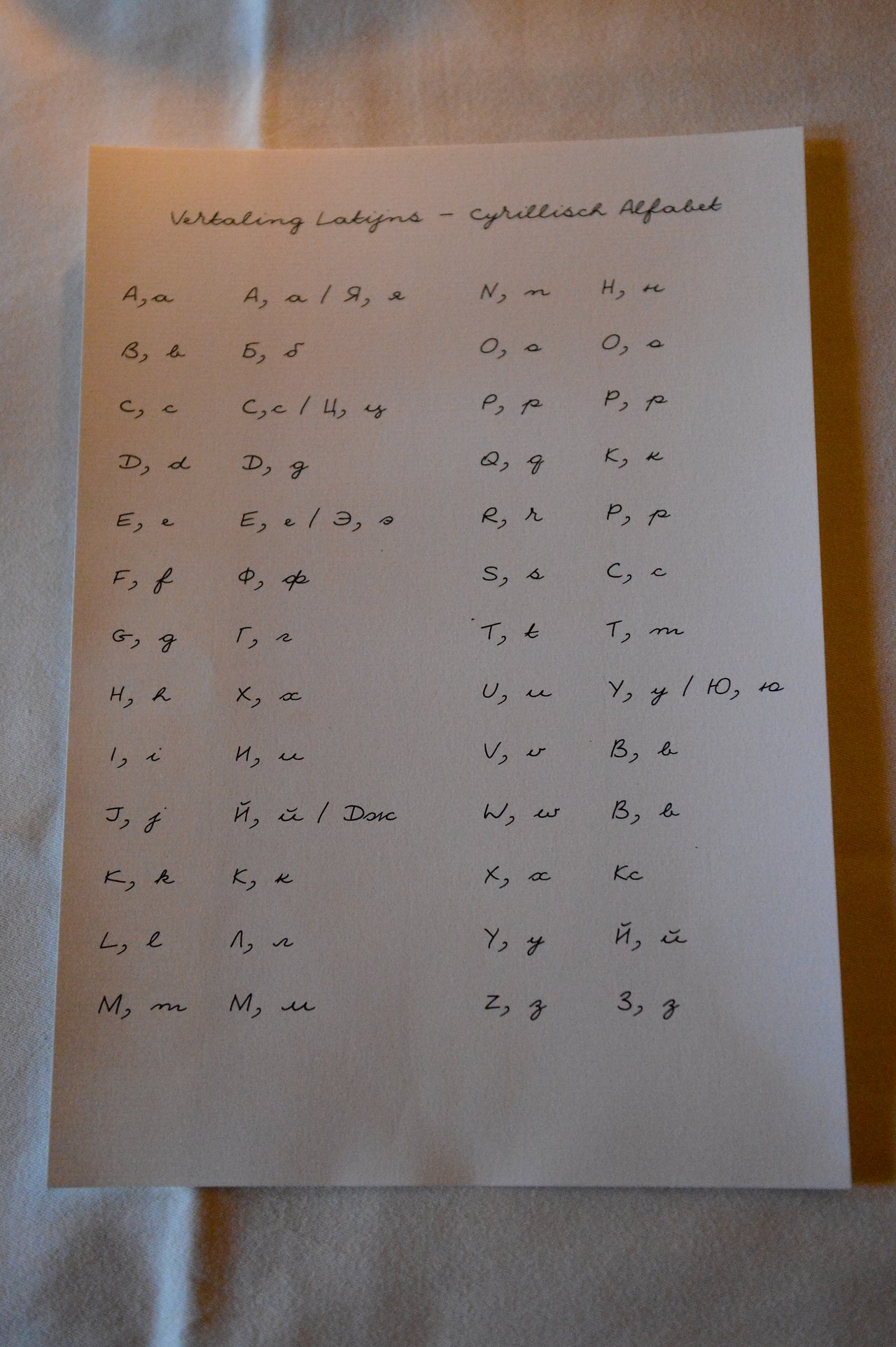 A handwritten chart comparing Latin and Cyrillic alphabets on a white sheet of paper placed on a table.