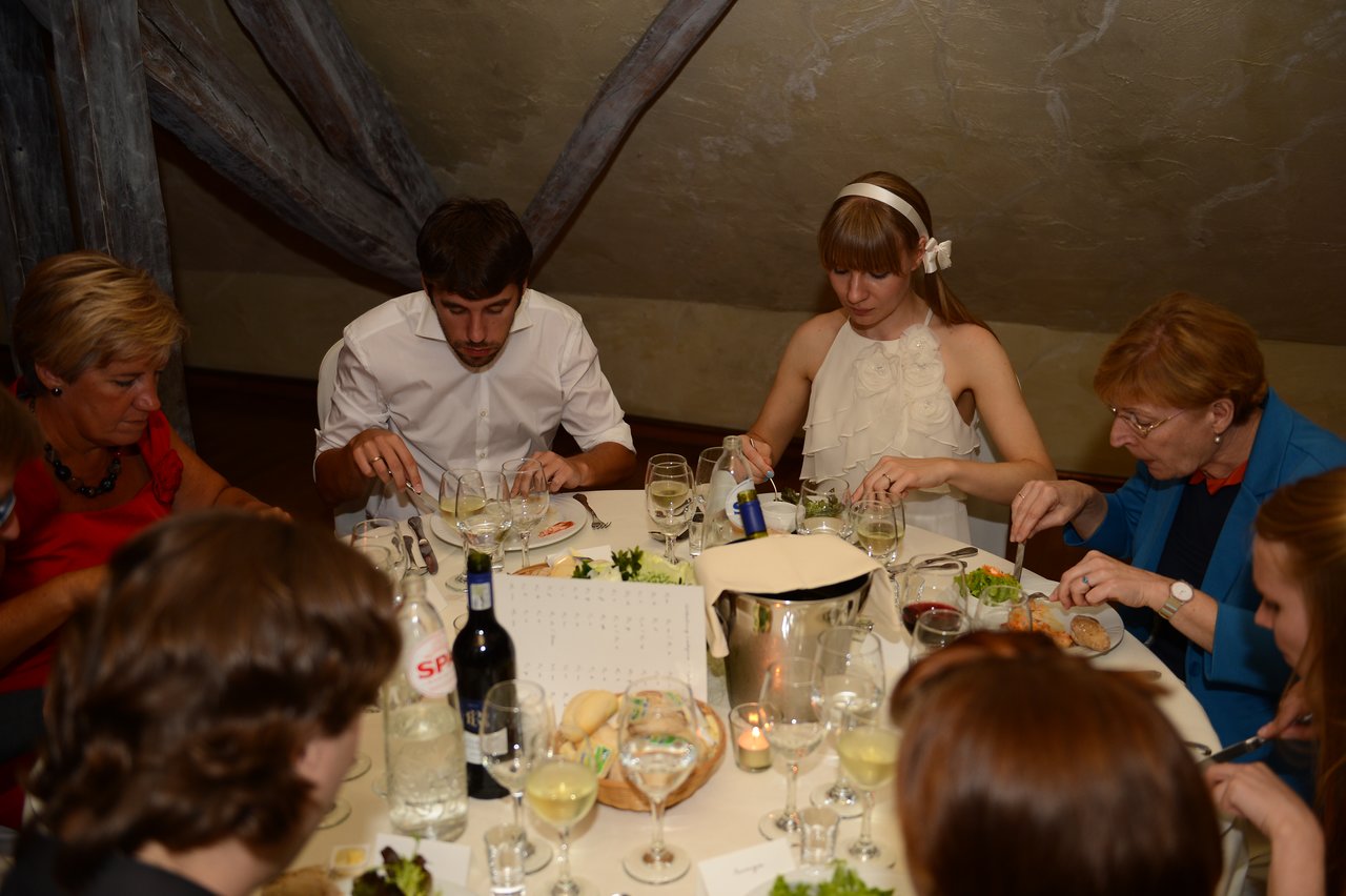 Wedding guests, including a bride in a white dress, are seated at a table, eating and enjoying a meal.