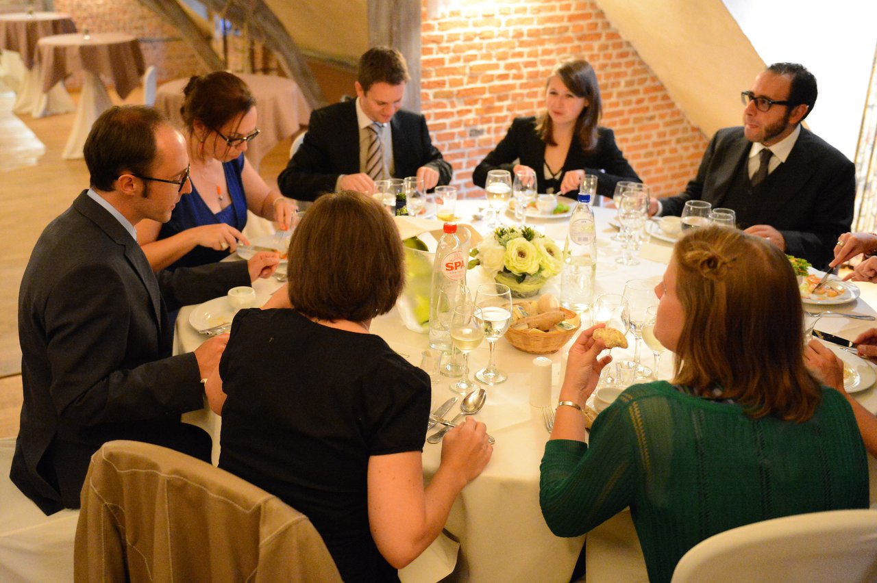 Guests in formal attire sit around a table, eating and talking during a wedding reception.