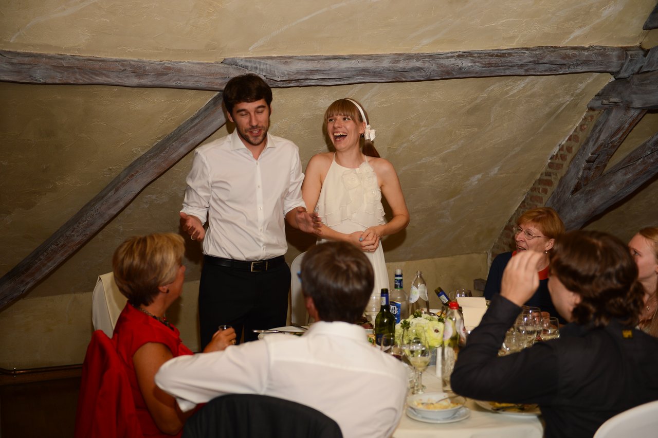 The bride and groom stand and smile while speaking to seated wedding guests at a dinner table.