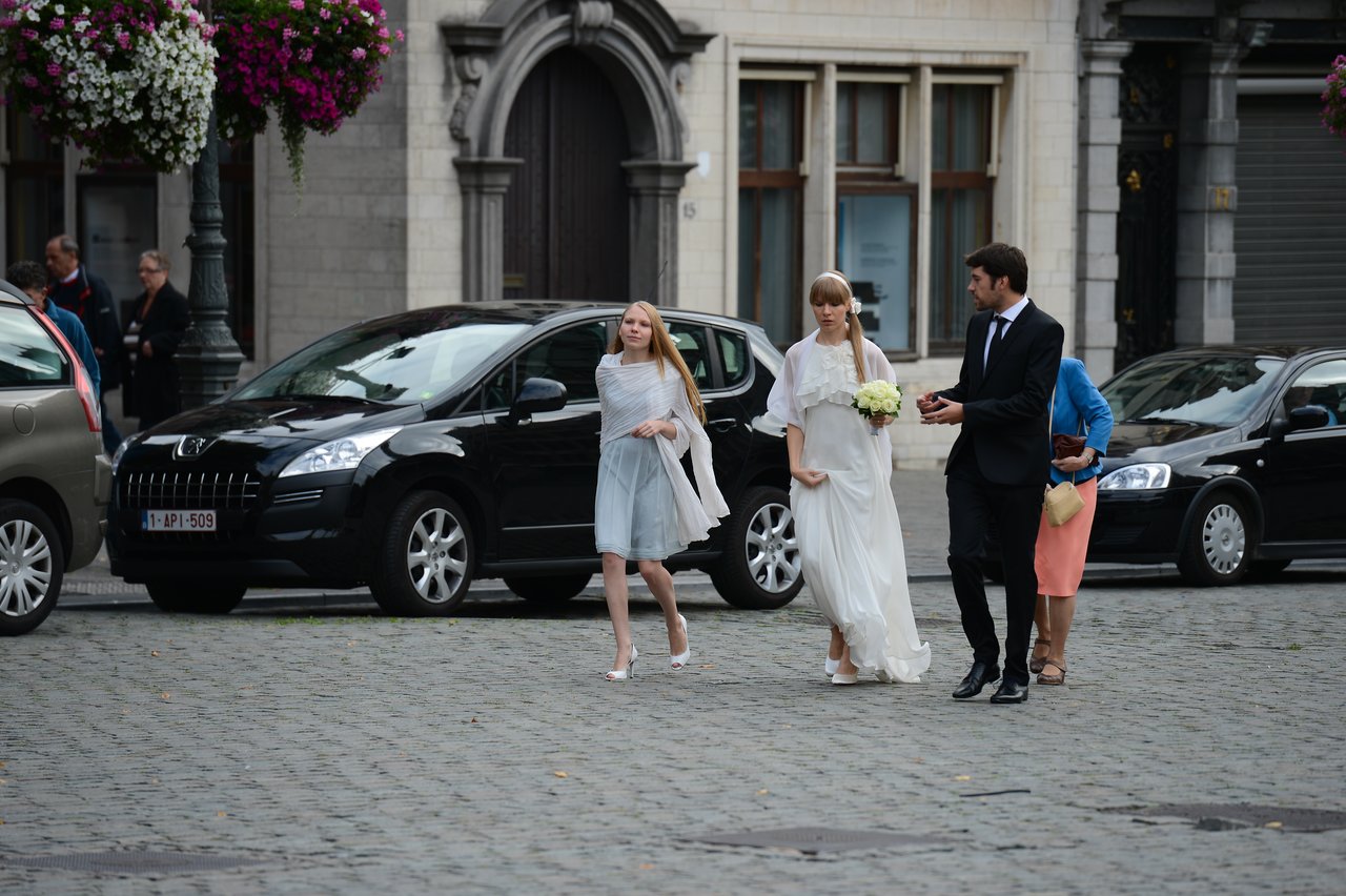 A bride in a white dress walks with a bouquet, accompanied by a groom and two guests on a cobblestone street.