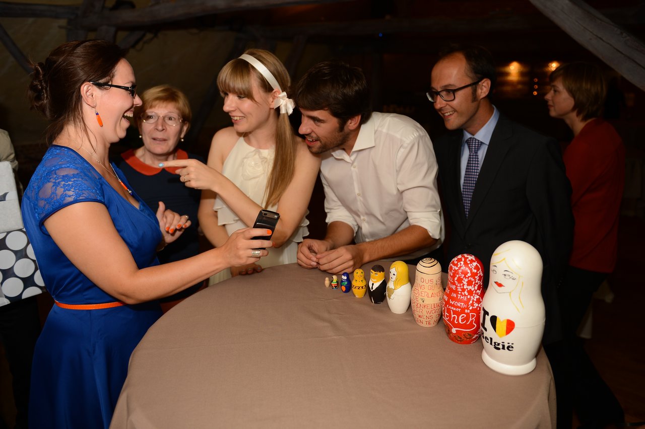 A group of people smiles and interacts around a table with decorated nesting dolls at a wedding reception.