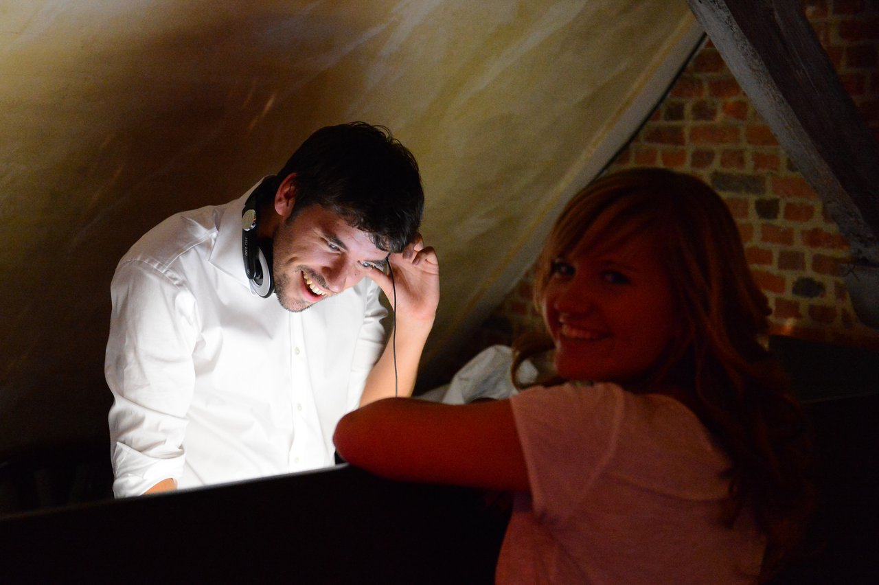 A smiling woman leans on a booth while a DJ in a white shirt adjusts his headphones and laughs.
