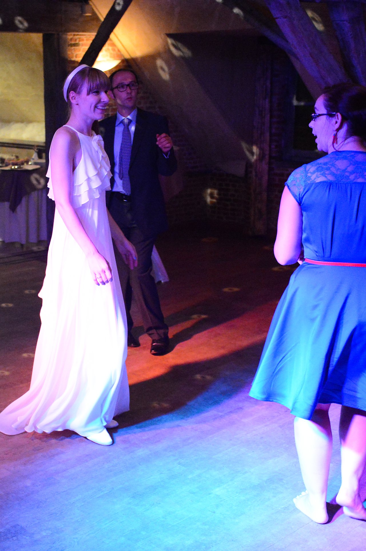 A bride in a white dress smiles while dancing with a guest in a blue dress at a wedding.