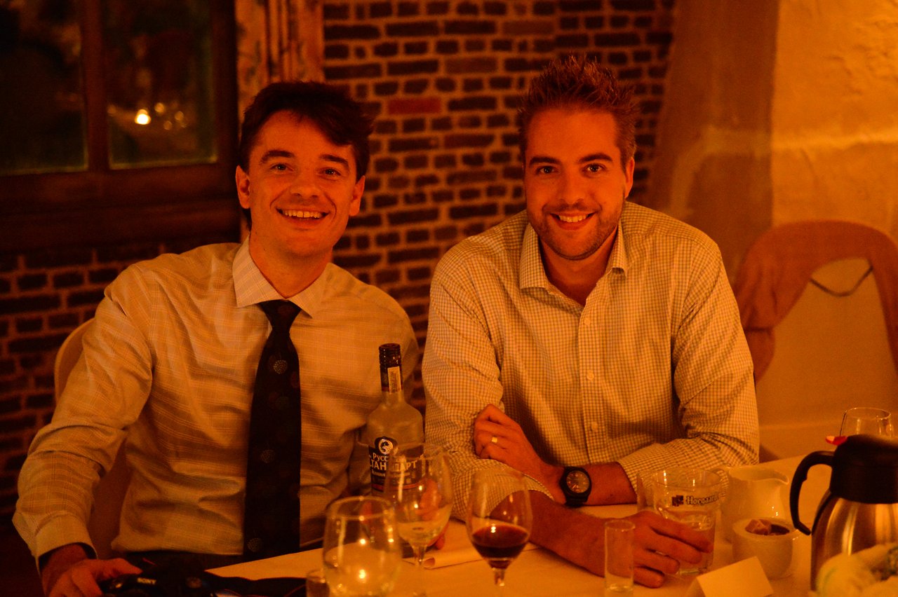 Two men sitting at a table, smiling at the camera during a wedding reception.